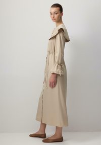 Abito maxi beige con colletto e polsini arricciati, vita stretta, bottoni frontali e texture liscia. Indossato con scarpe flats in rete marroni.