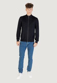 Hombre de pie vestido con una chaqueta negra de cremallera, vaqueros azules y zapatillas blancas, frente a un fondo liso claro.