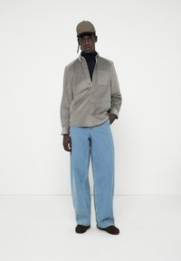 Camisa de algodón de pana gris con un bolsillo en el pecho, combinada con unos vaqueros de denim azul holgados. Unos zapatos marrones y una gorra con estampado completan el conjunto.