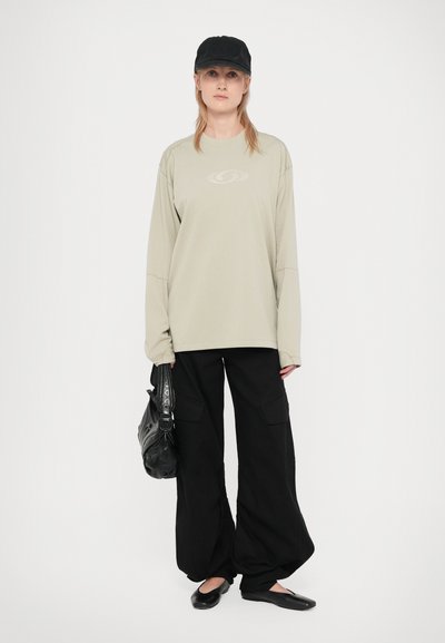 Camisa de manga longa, verde oliva claro, com uma textura subtil; calças cargo pretas soltas; sapatos pretos planos; boné preto e mala com detalhes em fita.