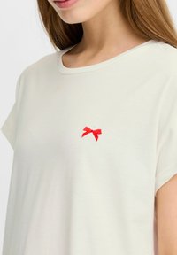 Kortærmet, cremefarvet t-shirt med en lille rød broderet sløjfe på venstre side af brystet. Blød, glat stoftekstur.