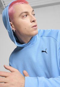 Sudadera azul clara con un bolsillo canguro en la parte frontal, hecha de tejido suave. Cuenta con un logo negro de Puma en el pecho y una capucha con cordón.