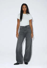 Frau mit langen schwarzen Haaren, die ein weißes kurzärmeliges Hemd, hoch taillierte weite graue Jeans und schwarze spitze Schuhe trägt, steht vor weißem Hintergrund.