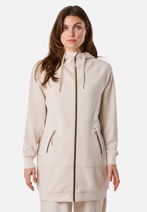Mujer que lleva una chaqueta con capucha beige con cremallera, mangas largas y dos bolsillos delanteros con cremallera, de pie delante de un fondo liso.