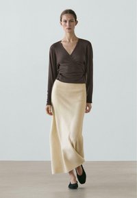 Braunes Wickeloberteil, beigefarbener knöchellanger Rock aus weichem Stoff, kombiniert mit schwarzen Slip-On-Schuhen. Schlichtes Design, figurbetonte Silhouette.