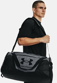 Black and gray duffel bag with an Under Armour logo, made from durable materials, featuring a wide strap and zip closure.