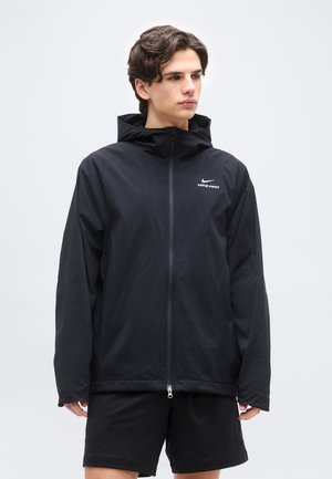 Jeune homme portant une veste à capuche noire Nike Pro avec une fermeture éclair frontale et un short noir, regardant sur le côté sur un fond uni.