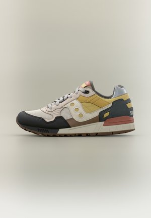 Nízký sportovní teniska v béžové, žluté, hnědé, černé a stříbrné barvě s tkaničkami a texturovanou podrážkou, zobrazená ze strany.