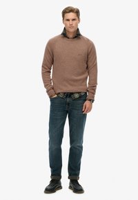 Bruine gebreide trui over een geruit shirt, blauwe jeans en zwarte veterschoenen. De trui heeft een ronde hals en geribbelde manchetten.