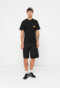 Μαύρο κοντομάνικο μπλουζάκι με τσέπη μπροστά και κίτρινη ετικέτα, συνδυασμένο με σκούρα τζιν σορτς και μαύρα αθλητικά παπούτσια με λευκές ρίγες.