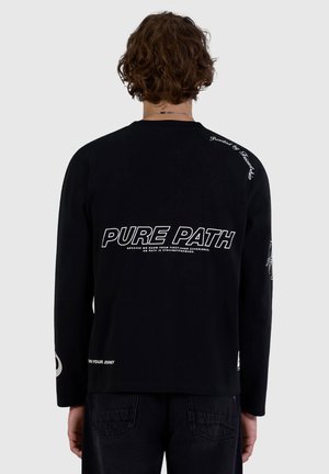Person with short curly hair facing away wearing a black long-sleeve shirt with white text "PURE PATH" on the back against a plain background.