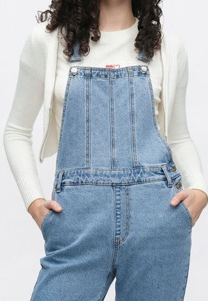 Vrouw met krullend haar die een lichtblauwe spijkerbroek overall en een witte top met lange mouwen draagt, met haar handen in de zakken.