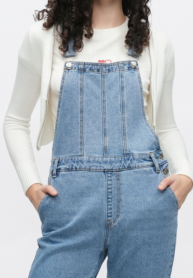 Femme aux cheveux bouclés portant une salopette en jean bleu clair et un haut blanc à manches longues, les mains dans les poches.