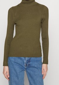 Olivgrüner, gerippter Rollkragenpullover mit langen Ärmeln, der eine taillierte Passform und ein strukturiertes Muster aufweist. Kombiniert mit blauen Jeans.