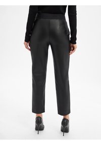 Pantaloni in ecopelle nera con vita alta decorata con marchio in rilievo, silhouette aderente e lunghezza alla caviglia, abbinati a tacchi neri.