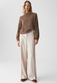 Brauner, flauschiger Strickpullover mit langen Ärmeln und runder Ausschnittform, kombiniert mit lockeren, hellbeigen, strukturierten Hosen. Braunes Schuhwerk.