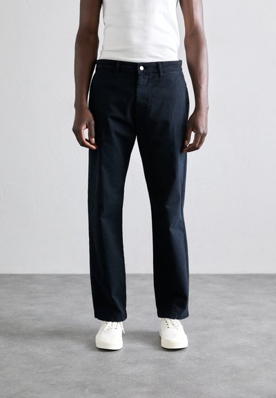 Mørkeblå bomuldsbukser med lige snit, med frontlukning med knap og sidelommer, båret med hvide sneakers.