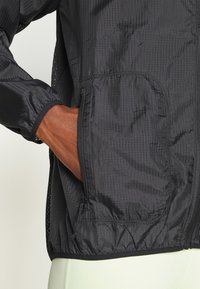 Cortavientos negro con superficie texturizada. Cuenta con puños elásticos, bolsillos laterales y dobladillo con cordón para un ajuste ajustable.