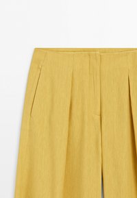 Gele broek met wijde pijpen, plooien aan de voorkant en zijzakken, getoond van de taille tot halverwege het dijbeen op een effen witte achtergrond.