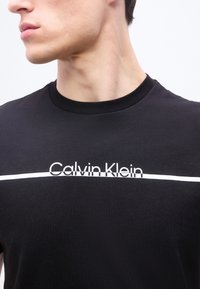 Νέος άνδρας που φοράει μαύρο μπλουζάκι με λευκή οριζόντια γραμμή και το κείμενο «Calvin Klein» στο κέντρο του στήθους.