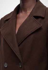 Gros plan sur une personne portant un manteau croisé marron foncé avec de larges revers et deux boutons visibles.
