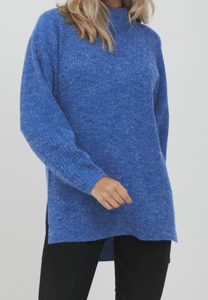 Femme portant un pull en maille bleu ample avec une texture côtelée et des fentes latérales, associé à un pantalon noir sur un fond uni.