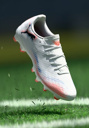 Chaussure de football blanche avec des accents dégradés bleus et roses, une tige texturée, six crampons roses et un logo noir sur le côté, flottant au-dessus de l'herbe.