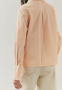 Chemise rayée orange clair avec des lignes verticales blanches. Dotée d'un col, de poignets boutonnés et d'un pli central dans le dos. Portée avec un pantalon beige.