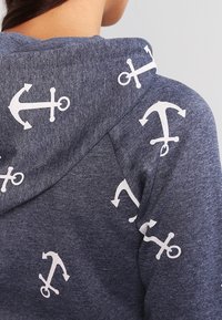 Felpa blu navy con una texture morbida, caratterizzata da motivi di ancore bianche sparsi sul tessuto. Il design include un cappuccio con cordini.