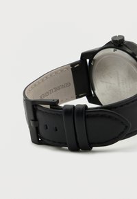 Črni usnjen pas za uro z gladko teksturo, nastavljivo sponko in srebrnim kovinskim pokrovom ohišja. Na notranji strani je vtisnjeno ime blagovne znamke.