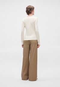 Άτομο με κοντά μαλλιά, ντυμένο με ένα εφαρμοστό κρεμ μπλούζα μακρύ μανίκι και χαλαρούς καφέ παντελόνες φαρδιάς γραμμής, που στέκεται μπροστά από ένα απλό φόντο.