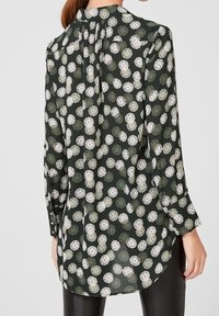 Blusa a maniche lunghe in tessuto verde scuro con motivo floreale bianco e pastello, caratterizzata da un retro plissettato e dettagli con bottoni sulle maniche.
