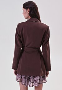 Bruine op maat gemaakte blazer met lange mouwen en een riem in de taille, gelaagd over een doorschijnende paisley rok in lichte paarse en bruine tinten.