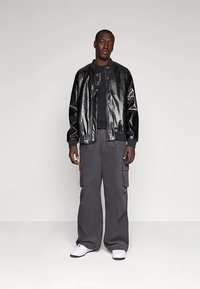 Svart bomberjacka med krokodilpräglat mönster över en svart tröja, kombinerad med grå cargobyxor och vita sneakers. Har texturerat material och lös passform.