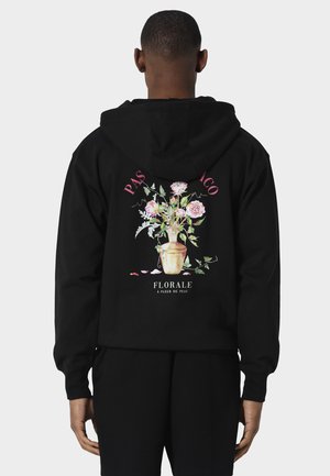 Hombre con sudadera negra con capucha con rosas rosas y hojas verdes en jarrón beige, texto "PAS DE CO" y "FLORALE A FLEUR DE PEAU" en la espalda.