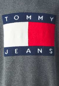 Maglione grigio con un grande logo composto da sezioni blu, bianche e rosse. "TOMMY" in testo bianco sopra "JEANS" su una banda blu scura. Texture morbida.