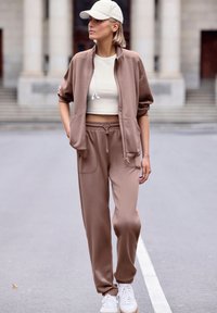 Hellbraune Zip-Jacke mit Taschen, kombiniert mit einem beigen Cropped-Top und passenden Jogginghosen, akzentuiert durch weiße Sneaker und eine beige Mütze.