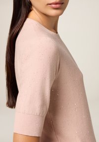 Kvinde vist fra skuldrene og ned iført en lyserød, kortærmet sweater med subtile glitrende prikker og langt, mørkt hår i en hestehale.