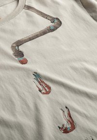 Katoenen T-shirt in lichtgrijs, met een grafiek van een bruine pijp met blauwe accenten en vallende figuren, gedrukt in subtiele, aardse kleuren.
