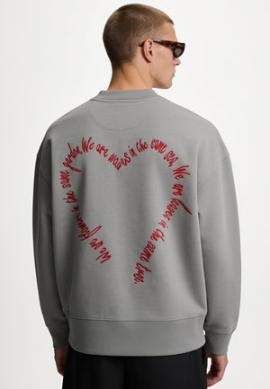 Mann trägt grauen Pullover mit rotem kursivem Text, der ein Herz auf dem Rücken bildet, und liest eine Botschaft über Fairness und Fürsorge.