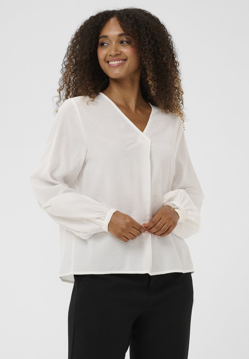 Femme aux cheveux bouclés portant un chemisier blanc à manches longues et un pantalon noir, souriante et regardant sur le côté sur un fond uni.