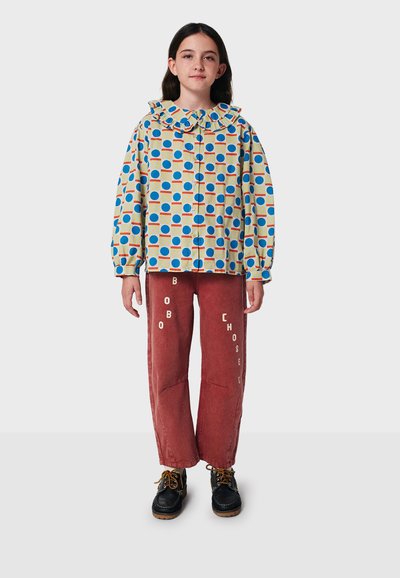 Camicia a maniche lunghe colorata con colletto arricciato, caratterizzata da un motivo geometrico di cerchi su uno sfondo chiaro; abbinata a pantaloni rossi e stivali neri.