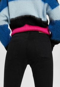 Jeans de mezclilla negra con dos bolsillos traseros, combinados con un suéter peludo multicolor que muestra rayas en azul, negro y rosa.