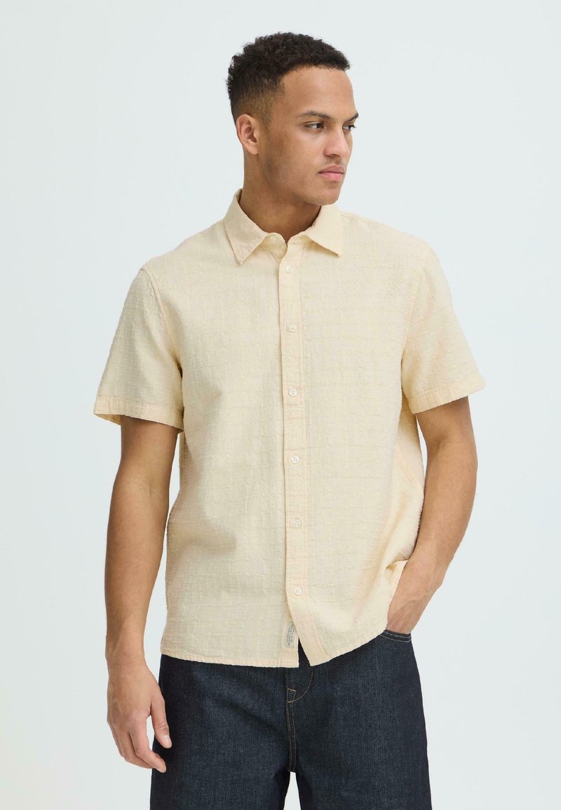 Jeune homme portant une chemise beige à manches courtes boutonnée et un jean bleu foncé, regardant vers sa gauche avec une main dans la poche.