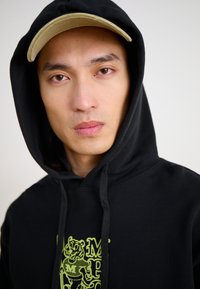 Black hoodie with a front graphic of a tiger in green. The model is wearing a light brown cap, with the hood pulled up around the face.