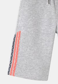 Světle šedé tepláky se dvěma svislými bočními pruhy v korálové a námořnické modři, s opakujícím se textovým vzorem "DOUCOU".