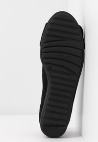 Semelle d'une chaussure noire avec des rainures, positionnée verticalement à l'angle d'un mur et d'un sol blancs.