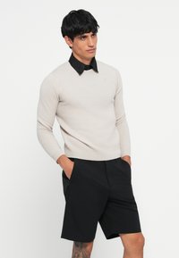 Sweater de lã bege com uma camisa preta de colarinho, apresentando um corte slim e punhos canelados, combinado com calções pretos e as mãos nos bolsos.