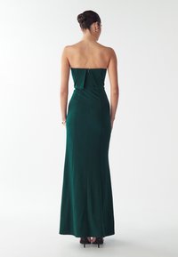 Naine, kelle juuksed on kokku pandud kuklasse, kannab rihmata tumeroheline põrandani ulatuv kleit ja musti kõrge kontsaga kingi, seistes ühtlase valge tausta ees.