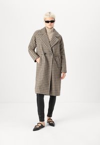 Smeđi i bež karirani kaput s velikim reverima, duplim dugmadi, bočnim džepovima, u kombinaciji s crnim kožnim hlačama i ravnim crnim cipelama.